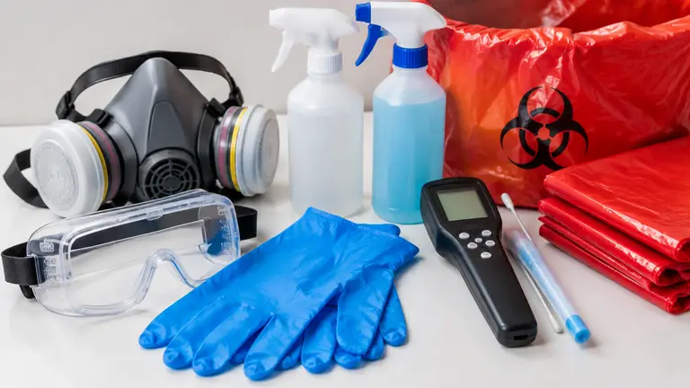 Respirator, goggles, gloves, disinfectant bottles, a surface test meter, and red biohazard bags laid out on a table