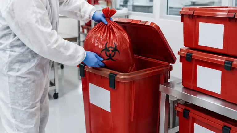 Gloved technician placing a sealed red biohazard bag into a rigid lidded waste container